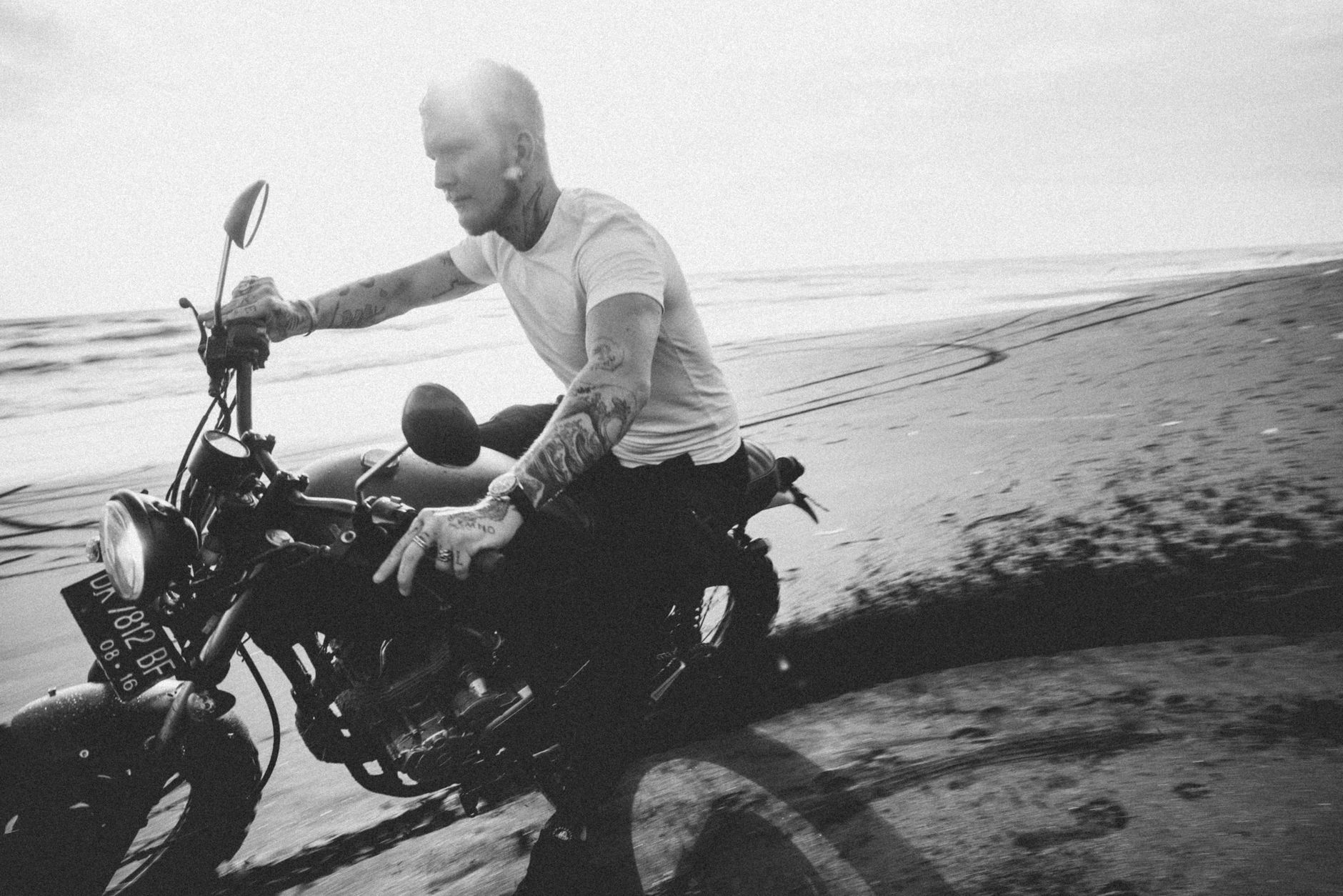 Brutal bearded hipster in t shirt and wristwatch riding motorbike with headlight on passing on sandy sea coast with trace in back lit