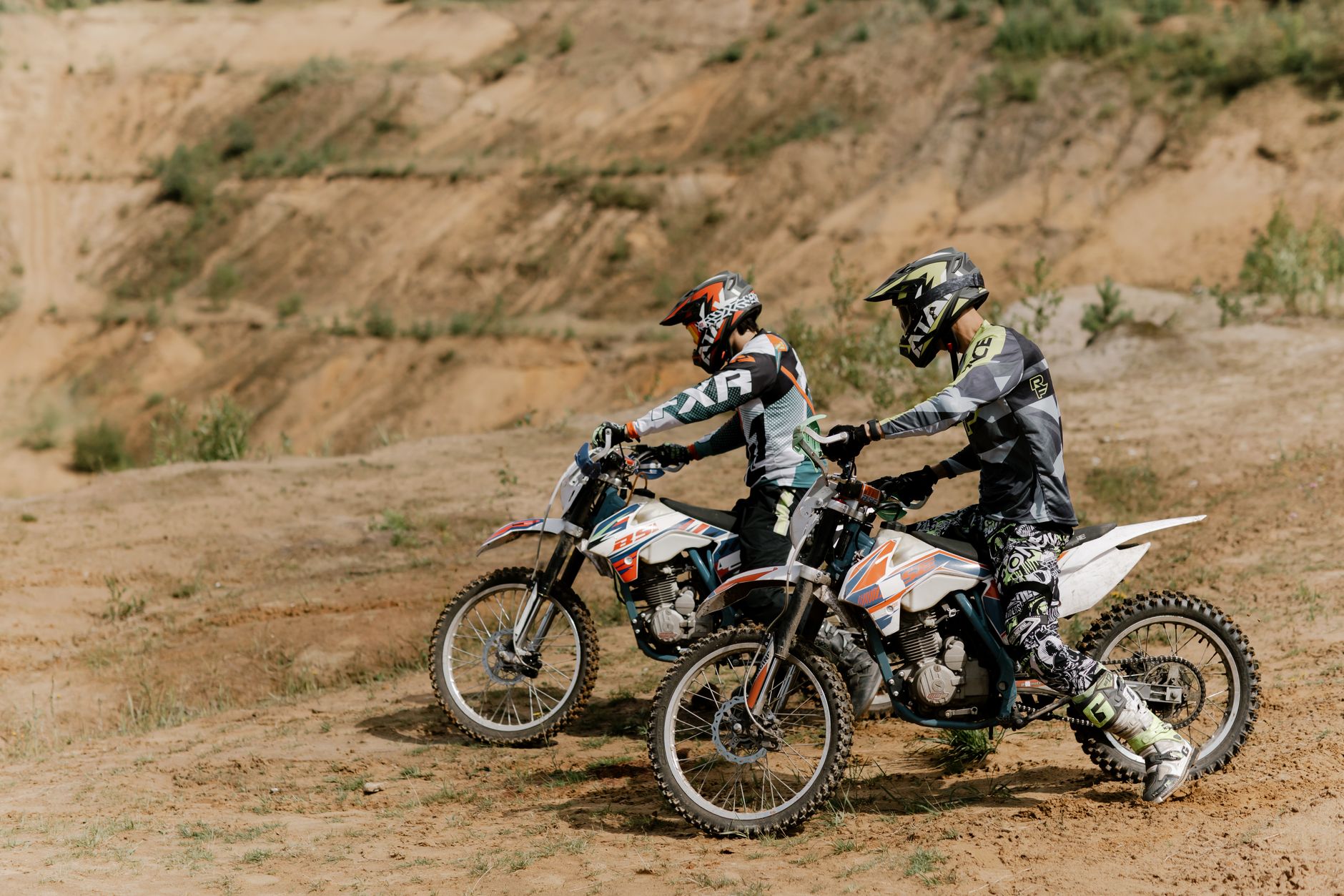 Two dirt bikers equipped with helmets prepare for a ride on a sandy outdoor trail. Enthusiastic and energetic.
